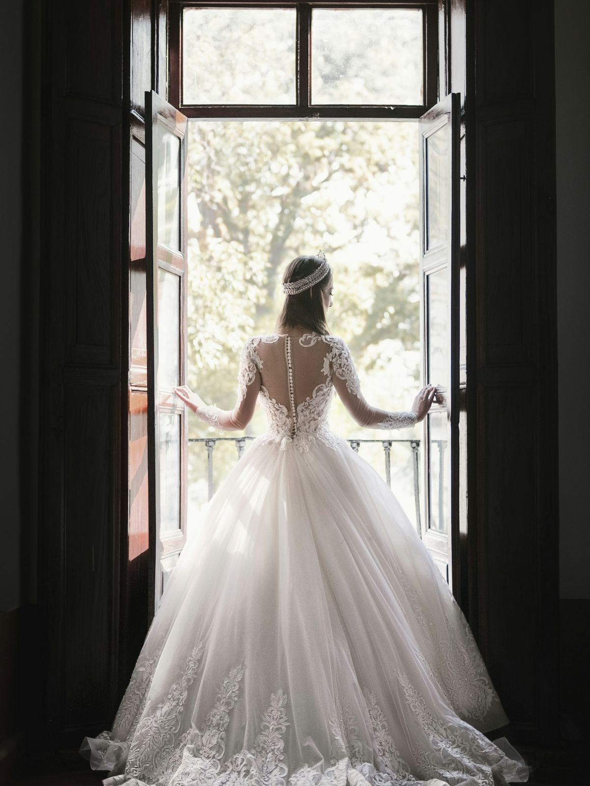 Bride in a lace-back ball gown standing at an open window